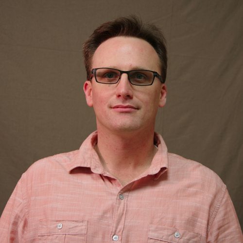 white man with short brown hair and glasses wearing a light coral button up shirt