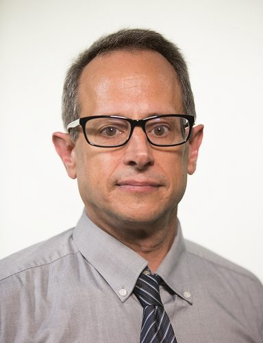 A white man with short hair and glasses wearing a grey button-up and grey and white striped tie.