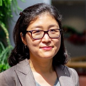 A medium-skinned woman with medium-length black hair wears glasses and a grey blazer while standing in front of a green houseplant and smiling.