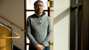 An Asian man with short, dark hair and glasses wears a grey sweater with 3/4 button closures inside of a building wiht large windows and a staircase behind him. He stands with his hands clasped in front of him.