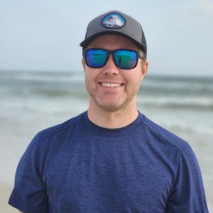 white man wearing a navy t-shirt, polarized sunglasses and a cap, standing in front of the ocean