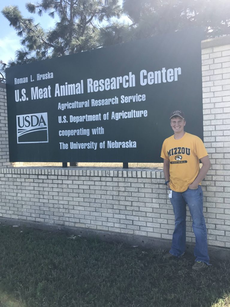 man in a gold mizzou shirt standing by a black USDA sign