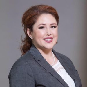 a light-skinned woman with redish-brown hair in a grey suit jacket and white blouse smiles at the camera.