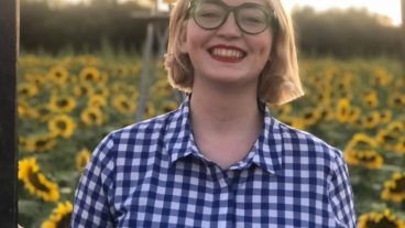 girl with short blonde hair and glasses in a blue and white gingham shirt standing in a field of sunflowers
