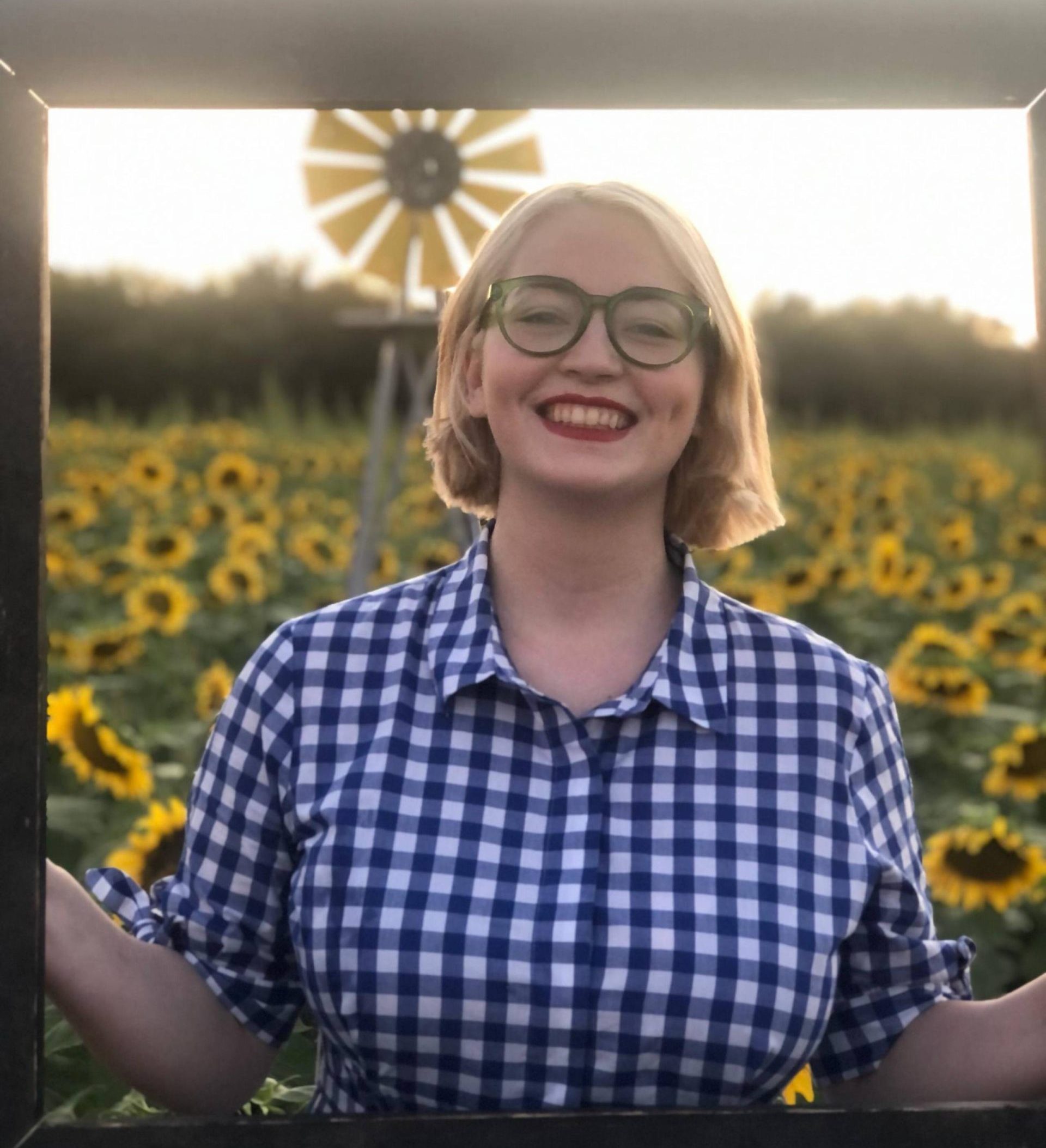 girl with short blonde hair and glasses in a blue and white gingham shirt standing in a field of sunflowers