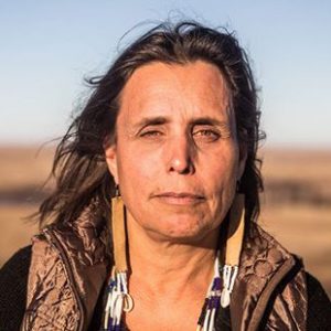 A medium-skinned woman with long, dark hair wearing a black shirt and brown vest with long earrings made from leather and feathers.