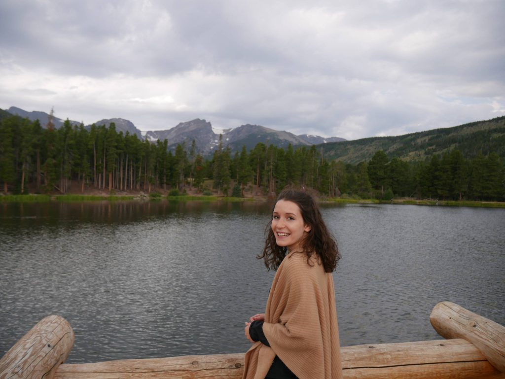 younger female with dark hair and a tan sweater is overlooking a lake with mountains in the background