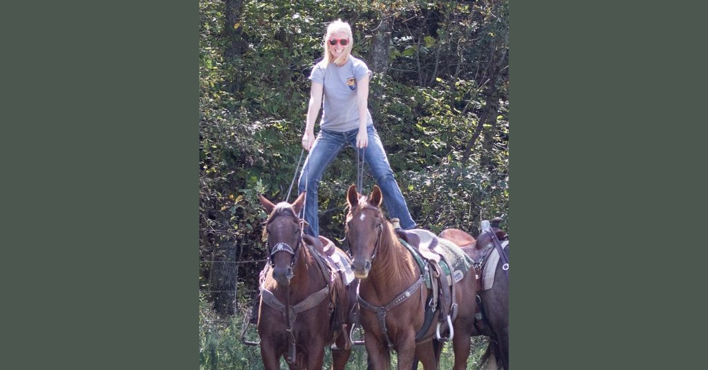 blonde female stands on top of a pair of horses