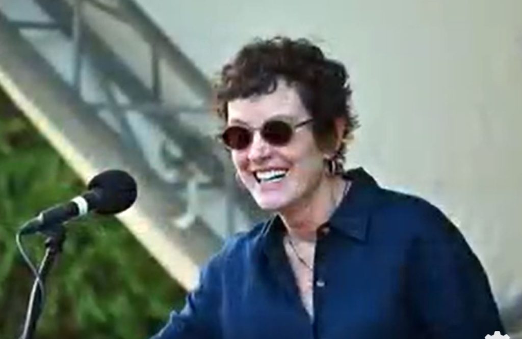 white woman with short brown curly hair, wearing a navy blouse and standing in front of a microphone