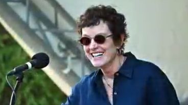 A woman in sunglasses smiles at a podium and talks into a microphone.