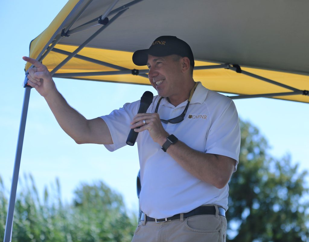 Dean Daubert wearing a white CAFNR polo and a black cap, standing under a pop up tent. He holds a microphone as he speaks to a crowd at a field day.