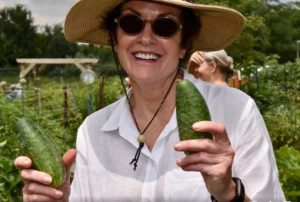 A woman wearing a white shirt and sunhat holds up a green vegetable and smiles.