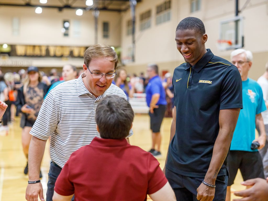 Coach Drinkwitz, left, white man in a striped polo and glasses stands beside Jalani WIlliams, black male student in a Mizzou polo, as they speak to a child on a basketball court.