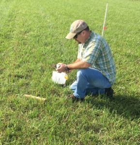 A man is bent down collecting samples from the grass and putting them in a container.
