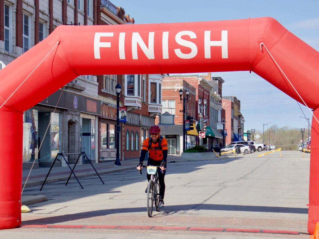a man on a bicycle wearing a red helmet and an orange shirt crosses a finish line on a downtown street