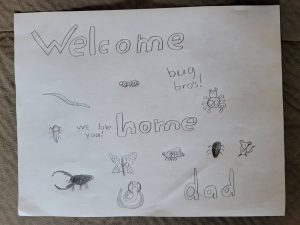 A white piece of paper with bubble letters reading "welcome home dad" and "bug bros!" and drawings of bugs.