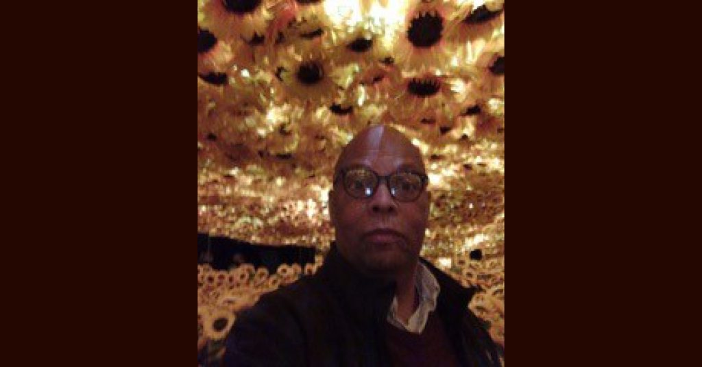 Black man with glasses wearing a dark coat inside a dimly-lit place