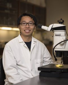 Peng poses for a portrait in a lab coat in front of his microscope. He is smiling and wearing glasses.