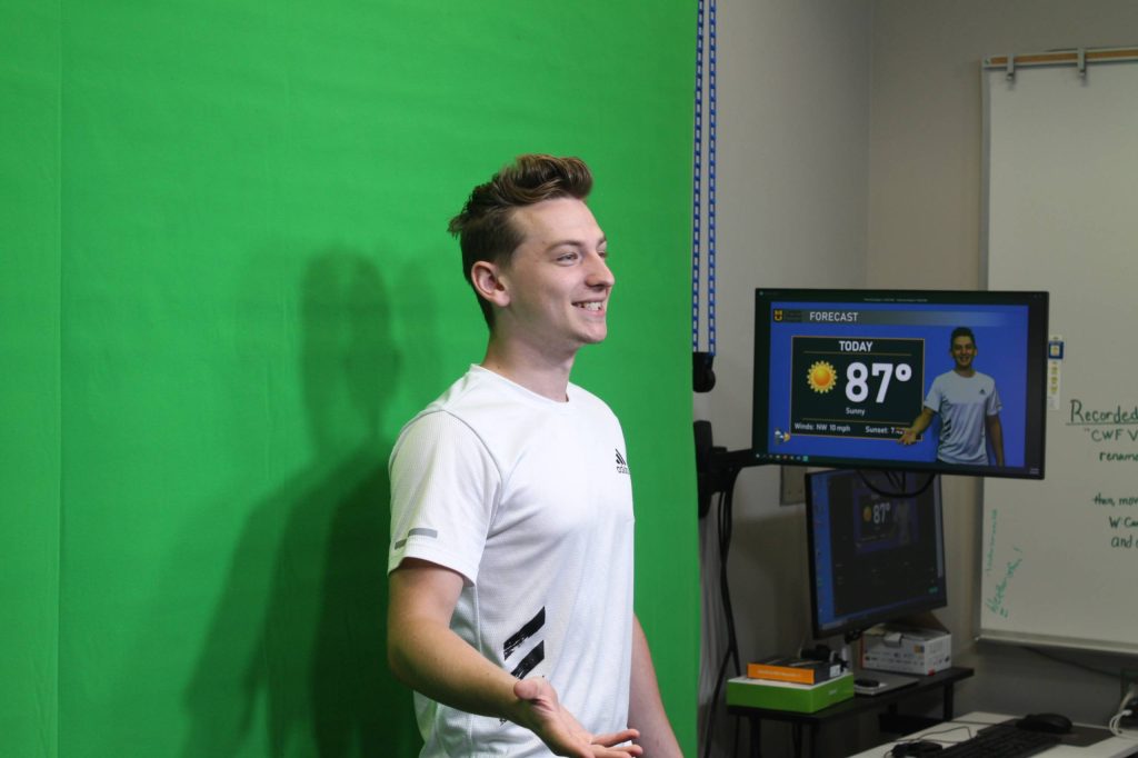 white male student with brown hair and wearing a white t-shirt standing in front of a green screen, recording a weather broadcast