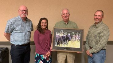 Rob Myers and Kelly Wilson are presented an award for the Center for Regenerative Agriculture by the Missouri Department of Conservation. Myers holds a framed photo of a grazing cow with an award plaque attached to the frame with Wilson and two MDC representatives flanking him.