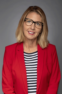 Annette Kendall, a muted blonde haired woman with glasses wearing a red blazer with a black and white striped shirt