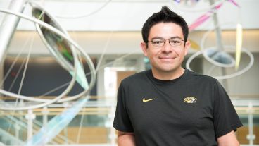 A man with dark hair, brown eyes, and black-rimmed glasses in a black t-shirt with a Mizzou athletic tiger emblem on it stands in front of a public art sculpture in the bond Life Sciences building. He is smiling and leaning on a railing