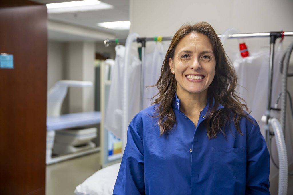 A white woman with long, brunette hair smiles while wearing a blue lab coat. Behind her are various medical equipment in a research laboratory.