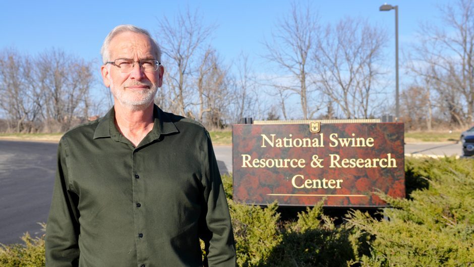 A white man with white hair and a white, neatly-trimmed beard in a dark grey button-up shirt stands in front of a sign that reads, "National Swine Resource & Research Center".