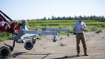 A white man in kaki pants and a white polot-style shirt with a white and grey ball cap stands next to a large piece of farm equipment with an electrocution bar mounted on front.