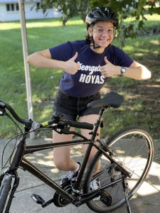 A young, brown-skinned woman wearing a blue Georgetown University t-shirt poses with a black bicycle while giving two thumbs up and smiling.