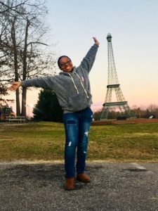 A young, black woman wearing a grey shirt and jeans stands with a joyous expression on her face and her arms outstretched with a replica of the Eiffel Tower in the background