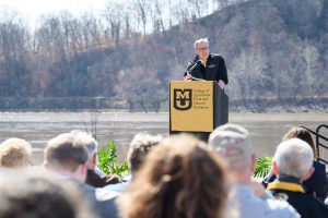 A white man with grey hair wearing a black vest wtih the CAFNR logo over a black sweter and gold tie speaks at an MU-branded podium with the Missouri River in the background. A crowd of people is in the foreground.