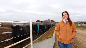 A woman stands outside next to her cattle operation. The sky is great and it looks windy.