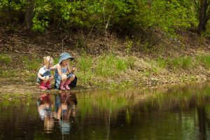 two blond, pre-school-aged girls sit beside a pond with fishing poles. They are wearing denim dresses and pink muck boots.