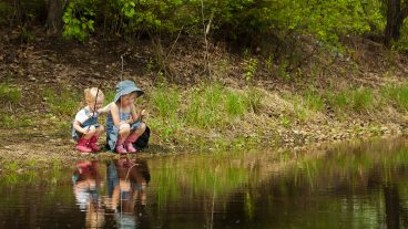 two blond, pre-school-aged girls sit beside a pond with fishing poles. They are wearing denim dresses and pink muck boots.
