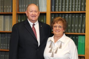 a man in a suit and red tie stands with a woman in a white top with bookshelves in the background