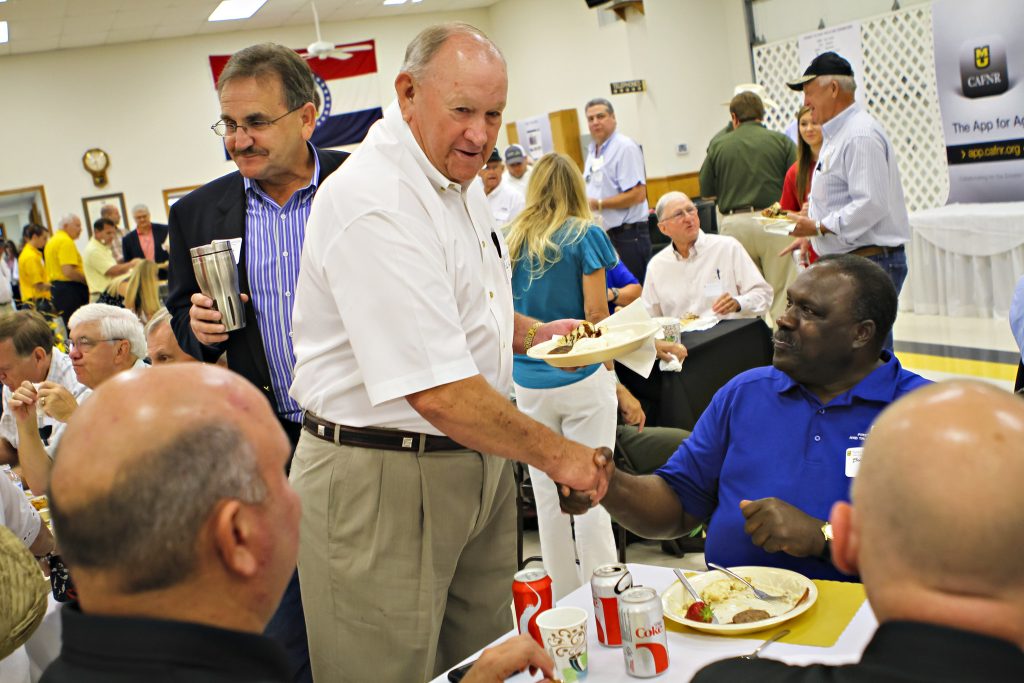An older, white man in a white button-up and kaki pants shakes hands with a black man in a blue polo while sitting at a table.