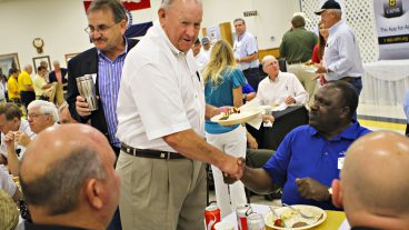 An older, white man in a white button-up and kaki pants shakes hands with a black man in a blue polo while sitting at a table.