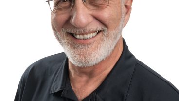 Headshot of Hank Stelzer. Man with white hair and glasses wearing a black and gold polo shirt