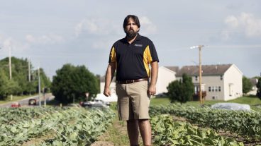 A man wearing a black and gold MU shirt stands in a row of crops.