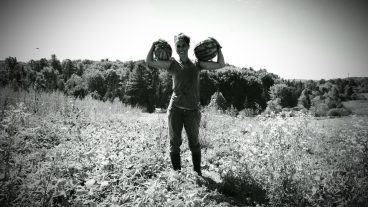 black and white photo of Kelly Wilson holding a watermelon on each shoulder, standing in the field