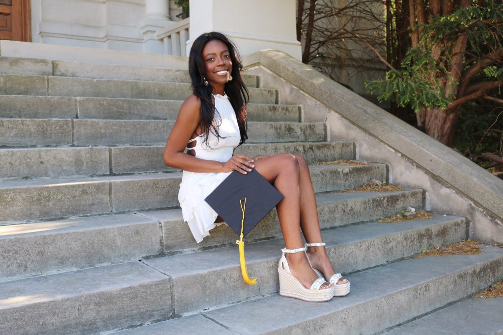photo of female student wearing a white dress and sandals, holding her grad cap, sitting on concrete steps
