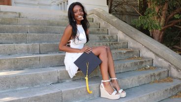 photo of female student wearing a white dress and sandals, holding her grad cap, sitting on concrete steps