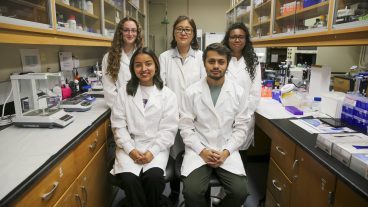 four women and one man all in white lab coats stand in a posed group amid shelves and counters of lab equipment