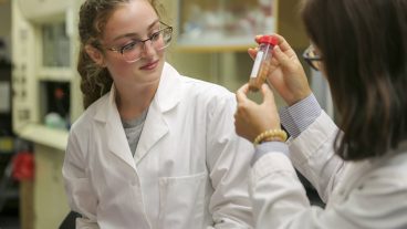 A white woman with curly hair and glasses looks at a test tube with a red cap that is being held by an asian woman with medium-length hair and glasses. Both are wearing white lab coats.
