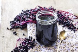 Canned elderberry jam surrounded by raw elderberries on a counter with a spoon.