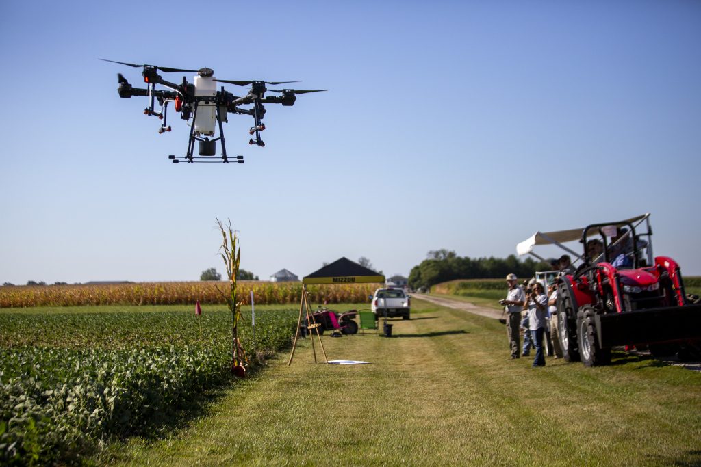 An agricultural drone flies above a crop of soybeans while people watch from a trailer with seats and an awning pulled by a red tractor.