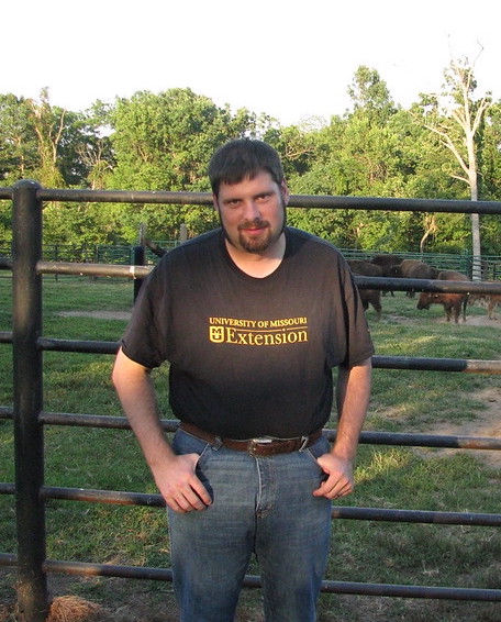 A man wearing a black MU Extension tshirt and jeans poses for a photo in front of a gate