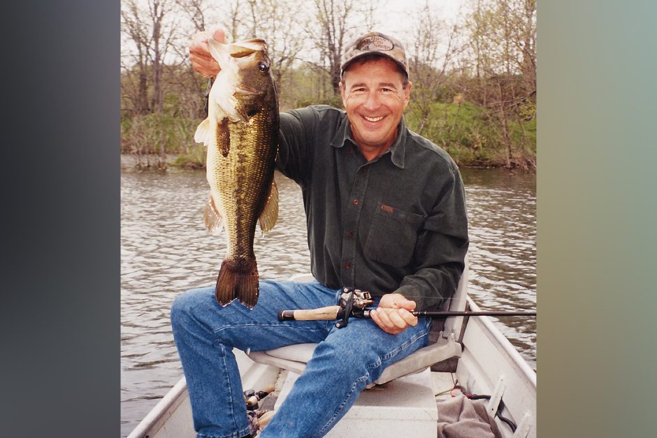 Johnny Morris wearing a ball cap and holding up a large mouth bass. He is sitting on a boat on a lake