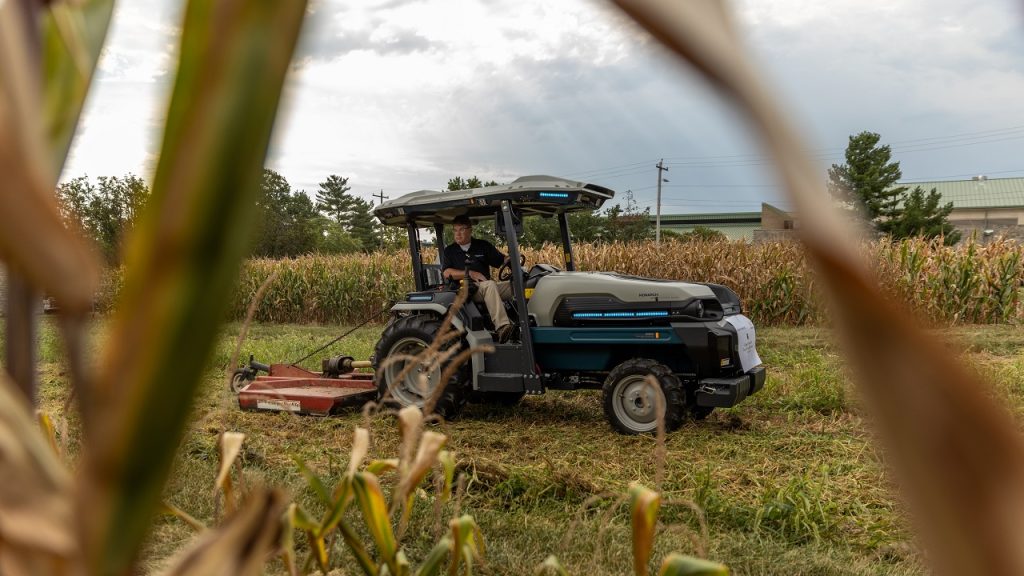 A light-skinned man in a black polo shirt and kakis drives a futuristic-looking tractor in a field. There are plots of corn behind the tractor and corn stalks in the foreground.